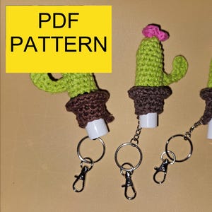 May include: Crocheted cactus-shaped hand sanitiser holders with keychains. The cacti are green with pink flower accents and brown pots. The image includes the text "PDF PATTERN" in a yellow box.
