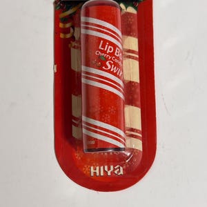 May include: Red lip balm with white diagonal stripes, labeled "Lip Balm Cherry Candy Swirl." The packaging is red with a candy cane design and the brand name "HIYA."