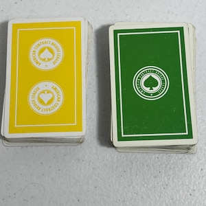 Può includere: Due mazzi di carte da gioco. Un mazzo ha il dorso giallo con la scritta "AMERICAN CONTRACT BRIDGE LEAGUE" e un simbolo di picche. L'altro mazzo ha il dorso verde con la stessa scritta e simbolo di picche. Entrambi i mazzi sono impilati.