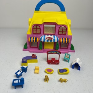 May include: A colorful plastic pet shop playset with a pink and yellow building, a blue handle, and various accessories. The set includes miniature tables, chairs, dog beds, and a dog show stand. The pet shop has a sign that says "Pet Shop".