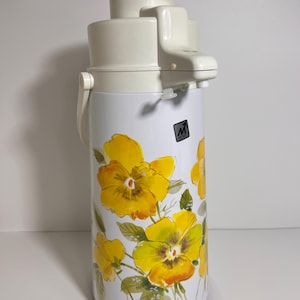 May include: A vintage white and yellow floral thermos. The thermos has a cream-colored top with a handle and a push-button dispenser. The body of the thermos is white with a pattern of yellow flowers and green leaves. A black and white logo is visible.