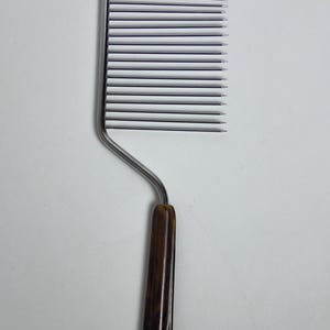 May include: A kitchen slicer tool featuring a rectangular head with fifteen parallel, thin, silver-colored metal tines. The tool has a bent metal neck connected to a smooth, dark brown, mottled tortoiseshell-patterned handle.