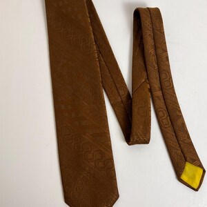 May include: A brown tie with a repeating paisley pattern. The tie is a solid brown color with a yellow lining at the tip. The tie is made of a textured fabric and is laying on a white surface.