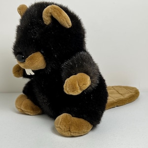 May include: A plush beaver toy with black fur and tan accents. The beaver has a tan snout, ears, and feet, with a flat, brown tail. It has two white teeth and black eyes. The toy is sitting upright.