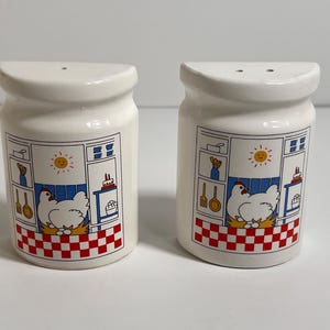 May include: A pair of white ceramic salt and pepper shakers. Each shaker features a kitchen scene with a hen, eggs, and a red and white checkered pattern. The design includes a sun, utensils, and a cake. The shakers have a curved top with two holes.