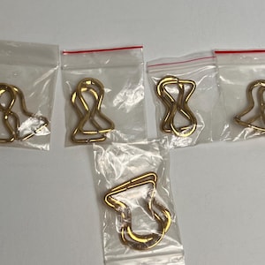 Könnte beinhalten: Fünf goldfarbene Metallverschlüsse in transparenten Plastiktüten mit roten Reißverschlussdichtungen. Die Verschlüsse haben ein einzigartiges, gebogenes Design und werden wahrscheinlich für Schmuck- oder Bastelprojekte verwendet. Die Beutel sind auf einer weißen Oberfläche angeordnet.