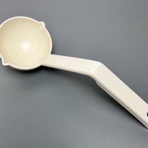 May include: Off-white plastic ladle with a round bowl and a long, angled handle. The handle has a hole at the end for hanging. The ladle is designed for scooping and serving liquids.