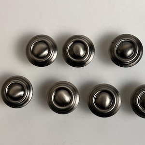 May include: A set of seven circular metal knobs with a brushed gunmetal finish. Each knob features a raised, smooth, rounded center dome surrounded by a concentric, recessed ring design, creating a layered, tiered appearance on the surface.