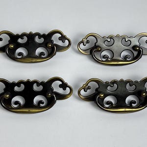 May include: Four matching drawer pulls made of dark, antiqued brass metal. Each handle features an ornate, openwork backplate with scrolling, curved cutouts and a central swinging bail handle attached by two small, rounded metal posts.