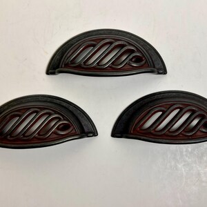 May include: Three semi-circular drawer pulls featuring a dark, weathered metal finish with a reddish-brown undertone. Each pull has an open, diagonal lattice design across the centre, creating a distinct arched shape for cabinet or furniture hardware.