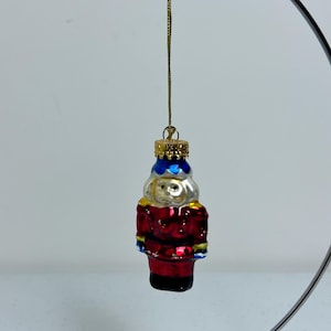 Puede incluir: Un adorno de vidrio rojo y plateado con forma de cascanueces. El adorno tiene un sombrero azul y dorado y está colgado de una cuerda dorada.