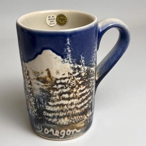 May include: Blue and cream ceramic mug with a mountain and forest scene. The mug features a dark blue rim and handle, with a landscape design in shades of brown and white. The words "Oregon" are written on the bottom of the mug.