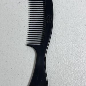 May include: A black plastic comb with fine teeth and a curved handle. The comb has a rounded top and a smooth, glossy finish. The handle is ergonomically designed for easy grip and use. A small logo is visible on the comb.