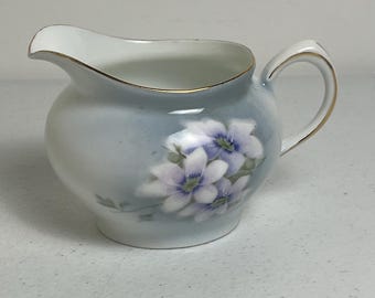 May include: A small, light blue creamer with a white interior and a gold rim. The creamer features a floral design with white and purple flowers. The handle and spout are also trimmed with gold.