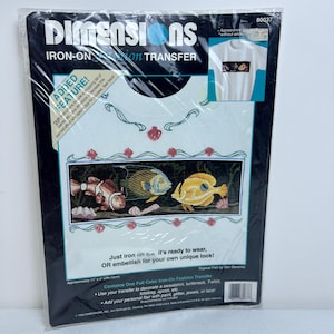 Puede incluir: Transferencia de planchado con un diseño colorido de tres peces nadando en un arrecife de coral. La transferencia está empaquetada en una bolsa de plástico transparente con fondo blanco y texto negro. El texto en el paquete dice "Dimensions Iron-On Fashion Transfer" y "Tropical Fish by Glam Geremia".