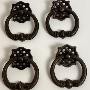 Peut inclure: Quatre poignées de tiroir en métal de couleur bronze, chacune dotée d'une poignée en anneau circulaire suspendue à une plaque arrière carrée décorative avec un motif ajouré en treillis et un petit détail de bouton central.