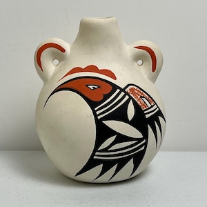 Pode incluir: Vaso de cerâmica creme com corpo arredondado, duas alças e gargalo estreito. O vaso apresenta um desenho estilizado de pássaro em preto e vermelho. O olho do pássaro é visível e o desenho inclui formas geométricas.