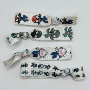 Op de afbeelding: Vier witte elastische haarbanden met herhaalde cartoonkarakterpatronen in blauw, rood, groen en roze. De haarbanden zijn geknoopt en klaar voor gebruik. Elke band is ongeveer 1 cm breed.