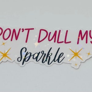 Op de afbeelding: Een witte sticker met de tekst "DON'T DULL MY SPARKLE" in rode en zwarte letters. De sticker is versierd met gouden glittersterren.