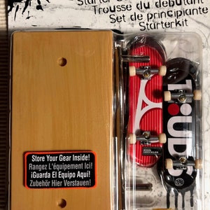 May include: Tech Deck Starter Set featuring two fingerboards, one red with white stripes and the text "Ryan Sheckler" and one black with white text "1. UDS". The fingerboards are displayed on a clear plastic backing with a wooden ramp.