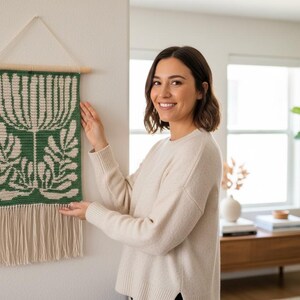 May include: A green and cream-colored woven wall hanging with a floral design. The rectangular piece is suspended from a wooden dowel and features a fringe at the bottom. The wall hanging is approximately 12 inches wide and 24 inches long.