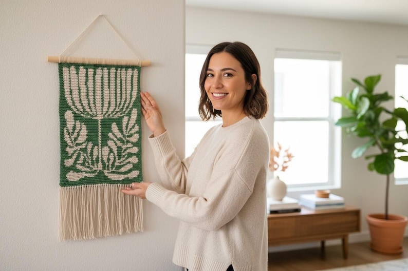 May include: A green and cream-colored woven wall hanging with a floral design. The rectangular piece is suspended from a wooden dowel and features a fringe at the bottom. The wall hanging is approximately 12 inches wide and 24 inches long.