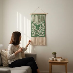 May include: A green and white woven wall hanging with a floral design. The tapestry is rectangular, with a wooden dowel at the top for hanging and a fringe at the bottom. The wall hanging is displayed on a white wall.