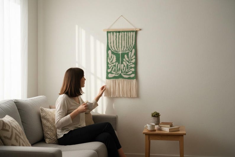 May include: A green and white woven wall hanging with a floral design. The tapestry is rectangular, with a wooden dowel at the top for hanging and a fringe at the bottom. The wall hanging is displayed on a white wall.
