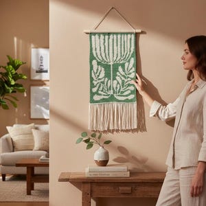 May include: A green and white woven wall hanging with a floral design. The tapestry is rectangular with a fringe at the bottom and is suspended from a wooden dowel with a leather cord. The wall hanging is a decorative home decor item.