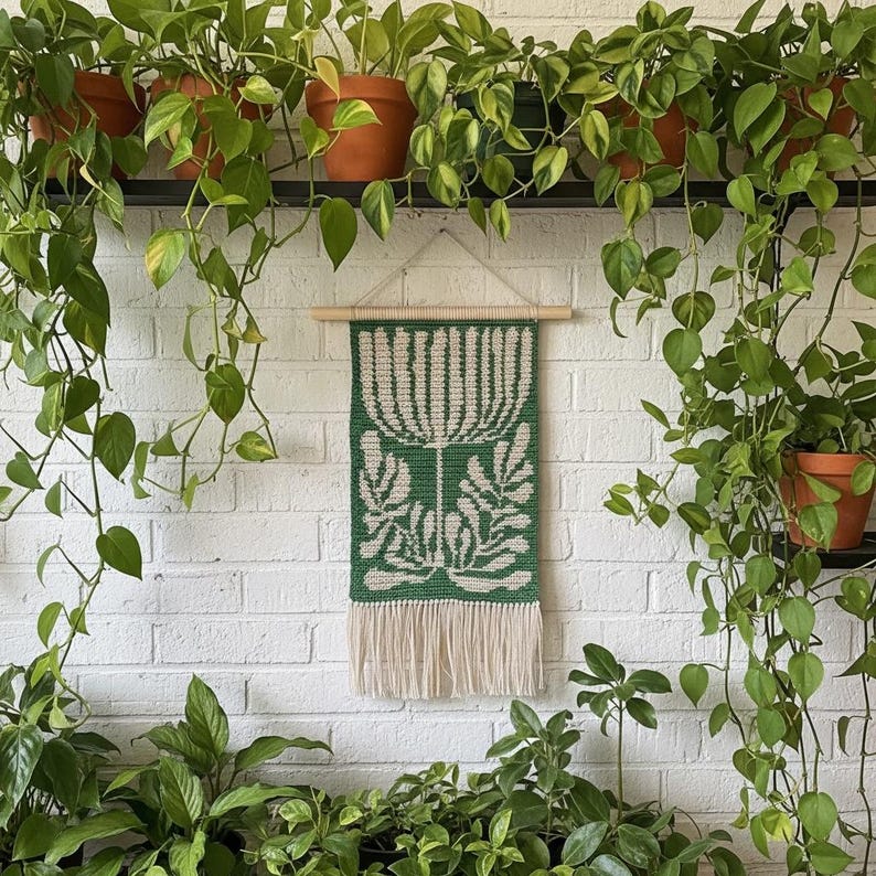 May include: A green and cream-colored woven wall hanging with a floral design. The rectangular tapestry features a central flower motif and leaf patterns, with a fringe at the bottom. It is suspended from a wooden dowel and hangs against a white brick wall, surrounded by potted plants.