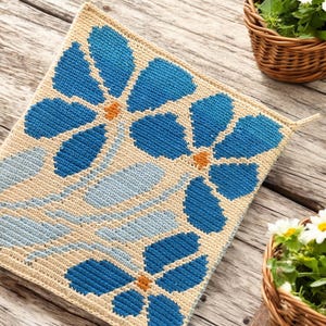Op de afbeelding: Een vierkant gehaakt vloerkleed met een bloemenpatroon. Het vloerkleed is beige met blauwe bloemen en oranje centra. De bloemen zijn gerangschikt in een herhalend patroon.