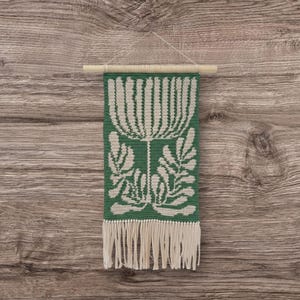 May include: A green and white woven wall hanging with a floral design and fringe. The rectangular tapestry features a large white flower and leaf pattern on a green background. It is suspended from a wooden dowel.