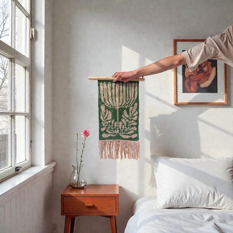 May include: A green and beige woven wall hanging with a floral design is held up by a wooden rod. The tapestry has a fringe at the bottom. A pink rose in a glass vase sits on a wooden nightstand.