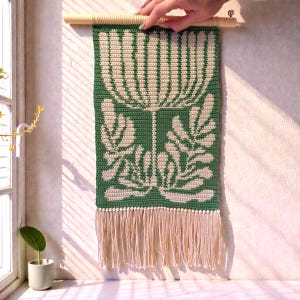May include: A green and cream-colored crochet wall hanging with a floral design. The piece is rectangular, with a cream-colored fringe at the bottom and a wooden dowel at the top for hanging. The floral pattern is a stylized botanical design.