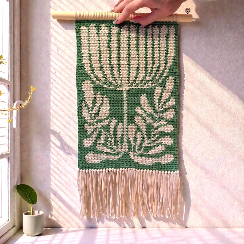 May include: A green and cream-colored crochet wall hanging with a floral design. The piece is rectangular, with a cream-colored fringe at the bottom and a wooden dowel at the top for hanging. The floral pattern is a stylized botanical design.