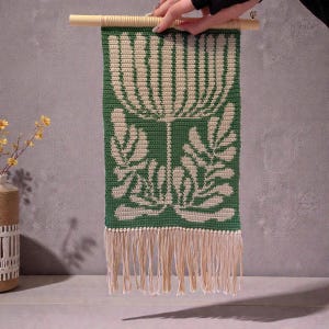 May include: A green and cream-colored macrame wall hanging with a floral design. The rectangular piece is suspended from a wooden dowel and features a fringed bottom. The design includes a large flower and leaf pattern.