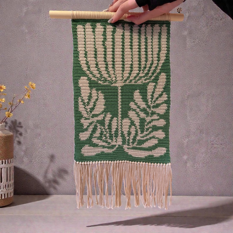 May include: A green and cream-colored macrame wall hanging with a floral design. The rectangular piece is suspended from a wooden dowel and features a fringed bottom. The design includes a large flower and leaf pattern.
