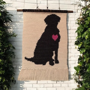 May include: A crocheted wall hanging featuring a black silhouette of a dog with a pink heart on its chest. The hanging has a fringe border and is made with beige yarn.