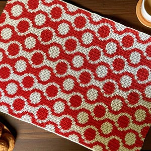 May include: A handmade crochet table runner featuring a repeating pattern of red circles on a cream background. The runner is rectangular and appears to be made of soft yarn. The design is a retro style.