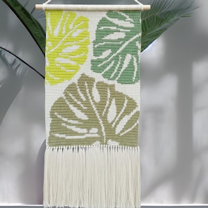 Può includere: Arazzo da parete tessuto con tre grandi foglie di monstera nei toni del verde e del giallo su sfondo bianco. Rifinito con frange nella parte inferiore e un'asta di legno nella parte superiore per appenderlo. Misura circa 60 cm di lunghezza.
