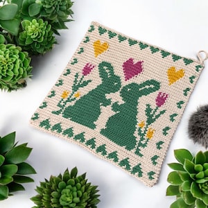May include: Cream-coloured crocheted potholder with a design of two green rabbits, purple and yellow flowers, and hearts. The potholder has a hanging loop and a decorative border. A charming, handmade kitchen accessory.