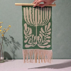 May include: A green and cream-colored crochet wall hanging with a floral design. The piece is rectangular, with a cream-colored floral pattern on a green background. It has a fringe at the bottom and is suspended from a wooden dowel.