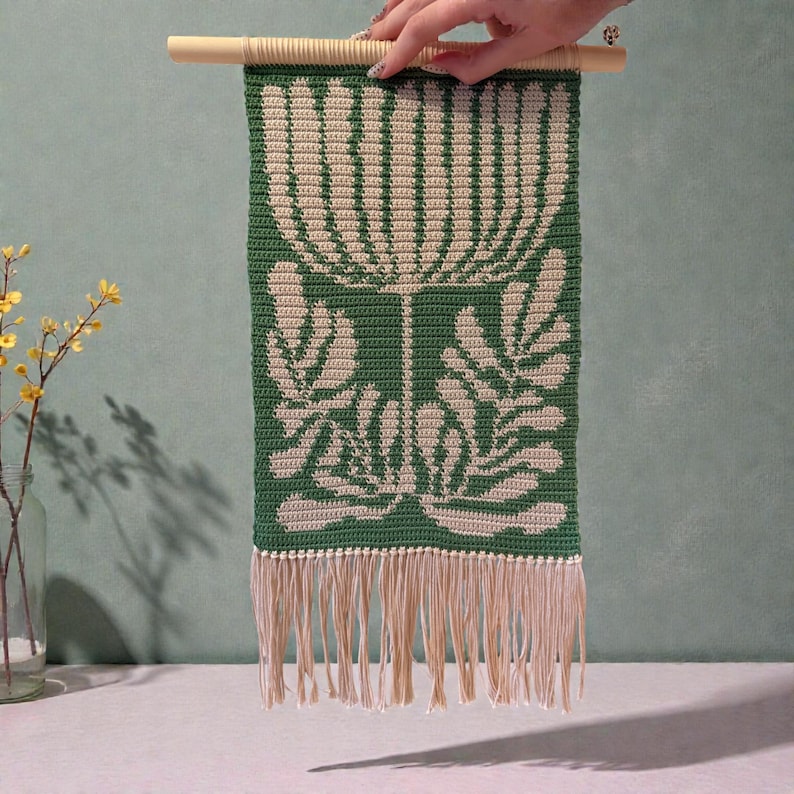 May include: A green and cream-colored crochet wall hanging with a floral design. The piece is rectangular, with a cream-colored floral pattern on a green background. It has a fringe at the bottom and is suspended from a wooden dowel.