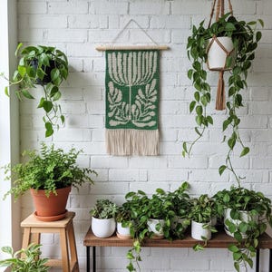May include: A green and cream-colored woven wall hanging with a botanical design is displayed on a white brick wall. Various potted plants, including ferns and trailing vines, are arranged on a wooden shelf and hanging from the ceiling, creating a natural, indoor aesthetic.