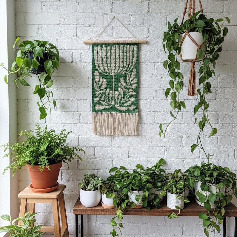 May include: A green and cream-colored woven wall hanging with a botanical design is displayed on a white brick wall. Various potted plants, including ferns and trailing vines, are arranged on a wooden shelf and hanging from the ceiling, creating a natural, indoor aesthetic.