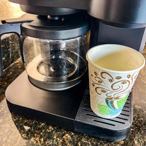 Op de afbeelding: Een zwarte koffiezetapparaat met een glazen kan en een papieren beker met een koffieontwerp. De beker staat op de lekbak. Het koffiezetapparaat staat op een gespikkelde aanrecht.
