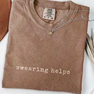May include: A brown Comfort Colors t-shirt with the phrase "swearing helps" in white lettering. A silver necklace with a small pendant rests on the shirt. A brown hat is partially visible in the upper left corner.