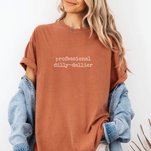May include: A rust-colored t-shirt with the text "professional dilly-dallier" in white. The shirt has rolled-up sleeves and is paired with a denim jacket. The overall style is casual and comfortable.