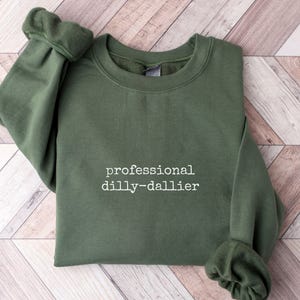 May include: An olive green sweatshirt with the phrase "professional dilly-dallier" printed in white. The sweatshirt is folded on a light brown wooden surface.