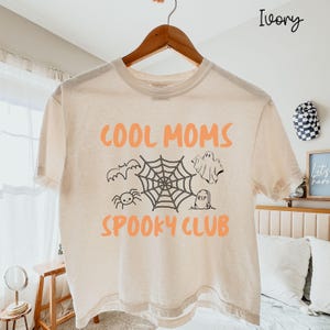 May include: An ivory short-sleeve t-shirt featuring the orange text "COOL MOMS SPOOKY CLUB" and a central black graphic illustration of a spider web, a bat, a spider, a ghost, and a tombstone with "RIP" written on it, all hanging on a wooden hanger.